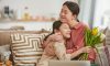 Asian girl with bunch of flowers hugging her happy mother sitting on sofa in living room, horizontal portrait