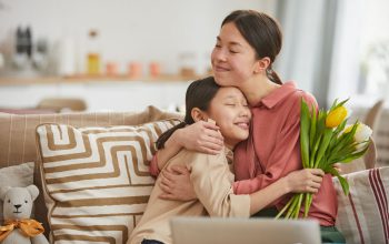 Asian girl with bunch of flowers hugging her happy mother sitting on sofa in living room, horizontal portrait