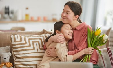 Asian girl with bunch of flowers hugging her happy mother sitting on sofa in living room, horizontal portrait