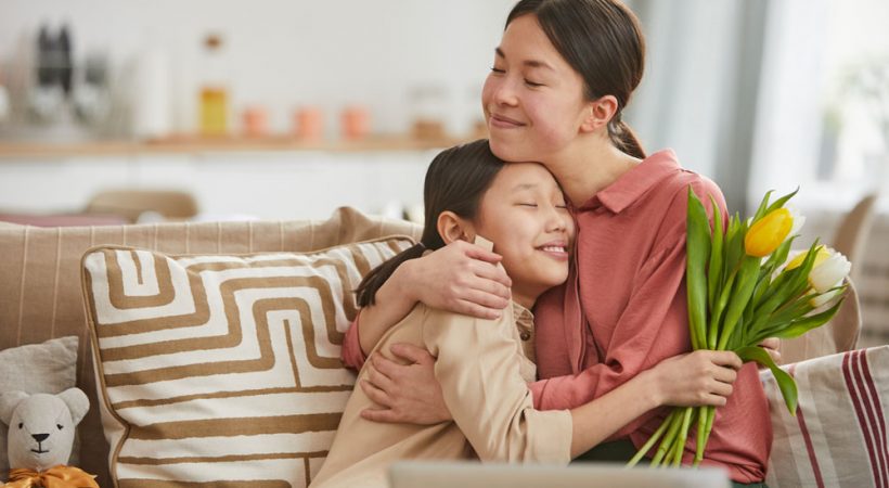 Asian girl with bunch of flowers hugging her happy mother sitting on sofa in living room, horizontal portrait